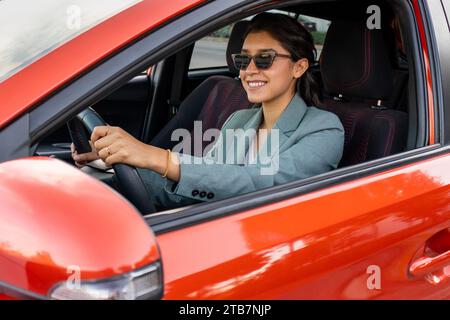 Fröhliche attraktive junge kaukasische Unternehmerin, die Sonnenbrille und Blazer trägt und lächelt, während sie ein modernes oranges Auto in der Stadt fährt Stockfoto