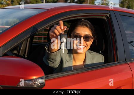 Porträt einer fröhlichen weißen schönen jungen Verkäuferin in Sonnenbrille lächelnd und mit Schlüssel, während sie während des Sonnenuntergangs in einem modernen orangen Auto sitzt Stockfoto