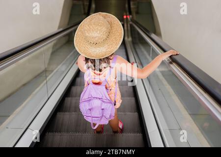 Hochwinkelvoller Körper eines anonymen Mädchens mit Strohhut und Fliederrucksack, der allein auf einer fahrenden Rolltreppe am Flughafen steht Stockfoto