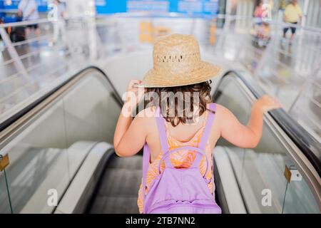 Hochwinkelvoller Körper eines anonymen Mädchens mit Strohhut und Fliederrucksack, der allein auf einer fahrenden Rolltreppe am Flughafen steht Stockfoto