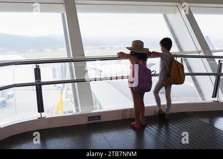 Rückansicht von nicht erkennbaren Geschwistern, die sich auf das Geländer lehnen und das Flugzeug auf der Landebahn vom Fenster aus betrachten, während sie zusammen in den Urlaub fahren Stockfoto