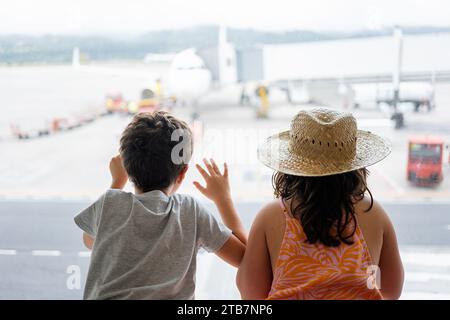 Rückansicht des anonymen Bruders und der Schwester in legeren Kleidern, die durch Fenster auf die Start- und Landebahn blicken, während sie auf die Abfahrt am modernen Terminal warten Stockfoto