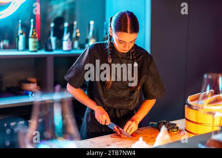 Fokussierte junge Küchenchefin in Uniform und mit langen geflochtenen Haaren schneidet Fleisch mit einem Messer auf der Theke, während sie eine Mahlzeit im Bistro zubereiten Stockfoto