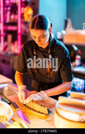 Selbstbewusste junge Küchenchefin mit langen Zöpfen in schwarzer Uniform und Handschuhen, die stehen, während sie frisch gekochtes Sushi in der Küche im Restaurant auf das Tablett legen Stockfoto