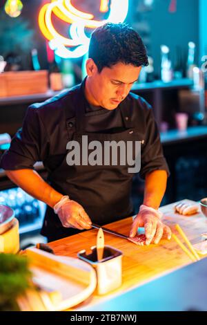 Fokussierter männlicher Koch in Handschuhen und schwarzer Uniform mit konzentriertem Fleischschnitt und Messer auf der Theke in der Küche in der modernen beleuchteten Sushi-Bar Stockfoto