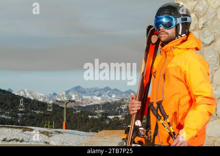 Ein Skifahrer in einer knallorangen Jacke steht mit seinen Skiern und blickt mit schneebedeckten Schweizer Alpen und Skiliften im Hintergrund in die Ferne Stockfoto