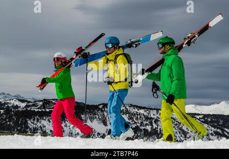 Drei Freunde, die in farbenfroher Skiausrüstung gekleidet sind und ihre Ski tragen, begeben sich freudig auf ein Winterabenteuer vor einer malerischen Bergkulisse. Stockfoto