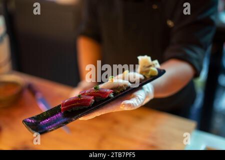 Ein Restaurantmitarbeiter präsentiert eine Sushi-Platte mit präziser Präzision, mit verschiedenen Nigiri auf einem traditionellen schwarzen Tablett. Stockfoto