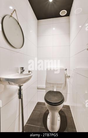 Innenausstattung eines kleinen gefliesten Badezimmers mit schwarzem Toilettensitz und Waschbecken in beleuchtetem, modernem Apartment Stockfoto