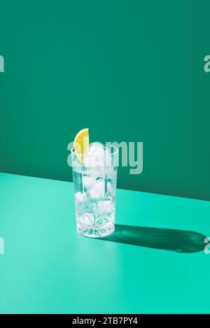 Ein mit Gin Tonic gefülltes Glas mit einer Limettenscheibe steht vor einem leuchtenden, grünen Hintergrund und wirft einen klaren Schatten. Stockfoto