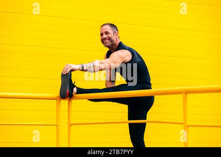 Ein lächelnder männlicher Athlet, der sein Bein auf dem Geländer streckt und wegblickt, während er in der Stadt gegen die gelbe Wand trainiert Stockfoto