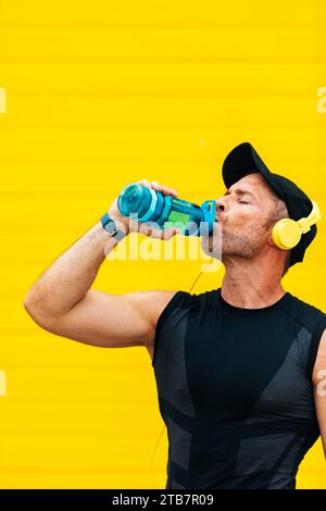 Männlicher Sportler in Sportkleidung, Trinkwasser aus der Flasche und Musik über Kopfhörer, während er während des Trainings gegen gelbe Wa Pause macht Stockfoto