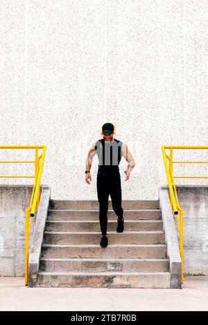 Ganzkörper eines aktiven männlichen Läufers in schwarzer Sportswear, der Musik über Kopfhörer hört und während des Trainings in der Stadt auf Betontreppen läuft Stockfoto