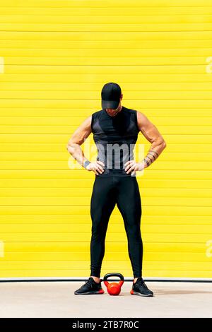 Anonymer muskulöser männlicher Athlet mit Armen akimbo, der mit Kettlebell auf der Straße über der gelben Wand steht, während er eine Pause vom Training hat Stockfoto