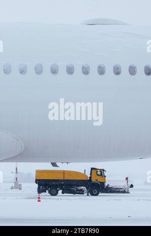 Winter frostiger Tag am Flughafen bei starkem Schneefall. Mit Schnee bedecktes Flugzeug gegen Schneepflüge, die die Start- und Landebahn des Flughafens räumen. Stockfoto