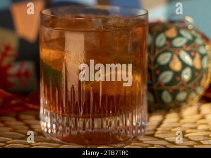 Tequila Negroni Cocktail in flachem Kugelglas mit Eis und Limette vor Weihnachtsdekoration mit Kugeln und rotem Band auf Goldmatte Stockfoto