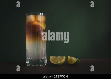 Dunkler und stürmischer Highball-Cocktail mit Eis- und Limettenkeilen auf Holztisch vor dunkelgrünem Hintergrund Stockfoto