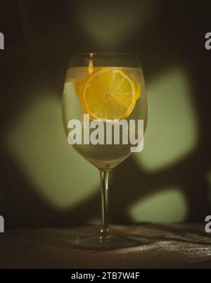 Gin-Tonic-Cocktail im Weinglas mit Eis- und Zitronenscheiben vor der hellgrünen Wand auf einem weißen Tischtuch Stockfoto