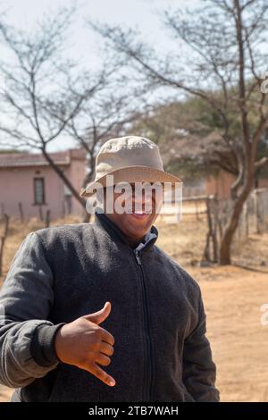 Ein afrikanischer Dorfmann mit einem Hut, der seine Hand hält, mit einem OK-Schild, einem Haus und einem Feldweg im Hintergrund Stockfoto