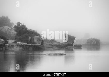 Ein Schwarzweiß-Foto mit einer idyllischen Landschaft mit einem See mit Felsformationen, die aus dem Nebel erscheinen Stockfoto