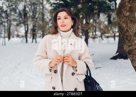 Modische junge Frau, die in einem schneebedeckten Winterpark mit weißem Lammfellmantel spaziert. Warme, stilvolle Kleidung Stockfoto