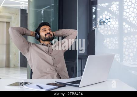 Geschäftsmann, der im Büro mit den Händen hinter dem Kopf und geschlossenen Augen stillte, Mann gut gemacht, erfolgreich ein Projekt mit Kopfhörern abgeschlossen Stockfoto