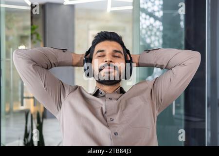 Geschäftsmann, der im Büro mit den Händen hinter dem Kopf und geschlossenen Augen stillte, Mann gut gemacht, erfolgreich ein Projekt mit Kopfhörern abgeschlossen Stockfoto