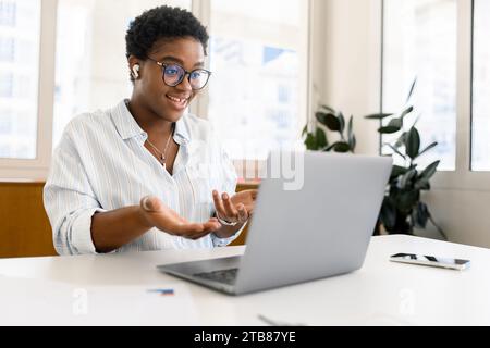 Freundlich lächelnde afroamerikanische junge Geschäftsfrau mit kurzer Frisur mit Laptop für Videoanrufe, Online-Gespräche und Gesten. Angenehme multirassische Angestellte, die Videoanrufe mit dem Kunden führt Stockfoto