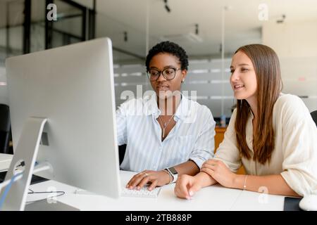 Zwei weibliche Kollegen diskutieren über das Projekt, sitzen nebeneinander im modernen Open Space Office, lächeln und beobachten gemeinsam am Computermonitor und genießen eine freundliche Arbeitsatmosphäre Stockfoto