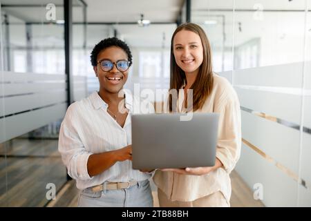 Zwei junge, glückliche weibliche Kollegen aus verschiedenen Rassen, die mit einem Laptop im Flur in einem modernen Büro oder einem Co-Working-Raum stehen und in die Kamera schauen. Vielseitiges Team-Konzept Stockfoto