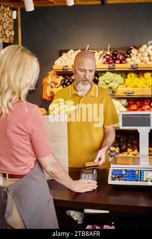 Konzentrierter bärtiger Kunde, der mit Kreditkarte für Früchte bezahlt, neben einem Reifen Verkäufer im Supermarkt Stockfoto