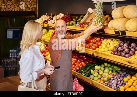 Fröhlicher bärtiger Verkäufer, der seiner Kundin neben dem Lebensmittelstand eine riesige Ananas zeigt Stockfoto