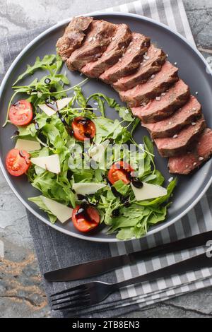 Italienischer Tagliata-Rindfleisch-Salat mit wilder Rakete, Kirschtomaten und Parmesankäse in Nahaufnahme auf einem Teller auf dem Tisch. Vertikale Draufsicht von oben Stockfoto