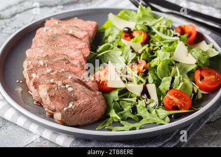 Gebratenes Steak Tagliata di Manzo, serviert mit Rucola, Kirschtomaten und Parmesan in Nahaufnahme auf einem Teller auf dem Tisch. Horizontal Stockfoto