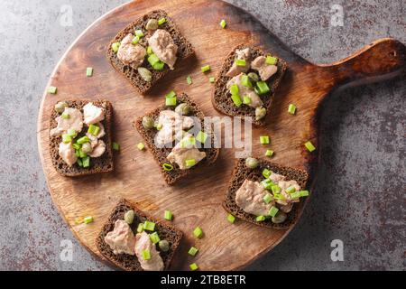 Hausgemachte Roggenbrotsandwiches mit Kabeljauleber, Kapern und grünen Zwiebeln in Nahaufnahme auf einem Schneidebrett auf dem Tisch. Horizontale Draufsicht von oben Stockfoto