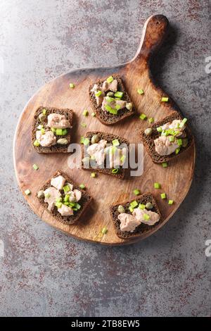 Sandwiches mit Kabeljauleber aus der Dose, grünen Zwiebeln und Kapern in Nahaufnahme auf einem Holzbrett auf dem Tisch. Vertikale Draufsicht von oben Stockfoto