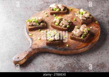 Gesundes Roggenbrot mit Dosenleber, Kapern und grünen Zwiebeln Nahaufnahme auf einem Schneidebrett auf dem Tisch. Horizontal Stockfoto