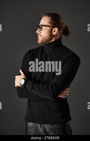 Bärtiger Mann in schwarzem Rollkragenpullover und Brille, der auf grauem Hintergrund steht und wegblickt Stockfoto