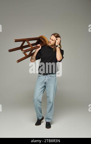 Die ganze Länge eines bärtigen Mannes mit langen Haaren, der in Jeans und T-Shirt posiert, während er einen hohen Hocker trägt Stockfoto