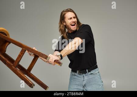 Emotionaler Mann mit langen Haaren in Jeans und T-Shirt mit hohem Hocker und grauem Schreien Stockfoto