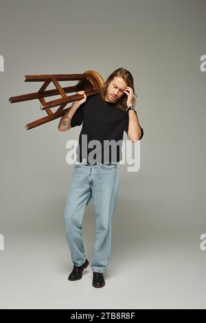 Die ganze Länge eines bärtigen Mannes mit langen Haaren, der in Jeans und T-Shirt posiert, während er einen hohen Hocker trägt Stockfoto