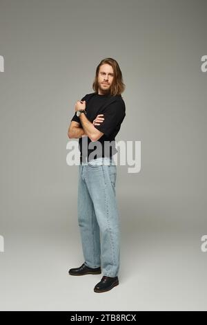 Die ganze Länge eines bärtigen und hübschen Mannes mit langen Haaren, der in Jeans und T-Shirt auf grauem Hintergrund posiert Stockfoto