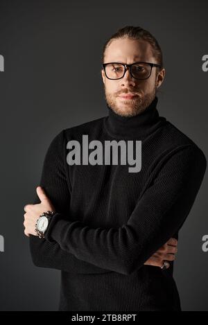 Bärtiger Mann in schwarzem Rollkragenpullover und Brille, der mit überkreuzten Armen auf grauem Hintergrund steht Stockfoto