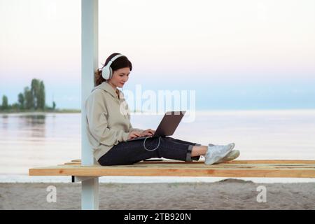 Junge freischaffende Frau, die am Laptop arbeitet und am Strand Kopfhörer trägt und auf einer Bank sitzt Stockfoto
