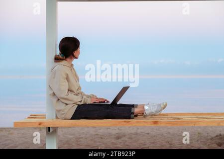 Junge, traurige, nachdenkliche freischaffende Frau, die auf einer Strandbank sitzt und auf den wunderschönen See und die Berge blickt Stockfoto