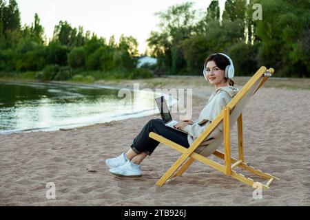 Glückliche, lächelnde Frau, die am Strand mit Laptop und Kopfhörern auf einer Liege sitzt Stockfoto