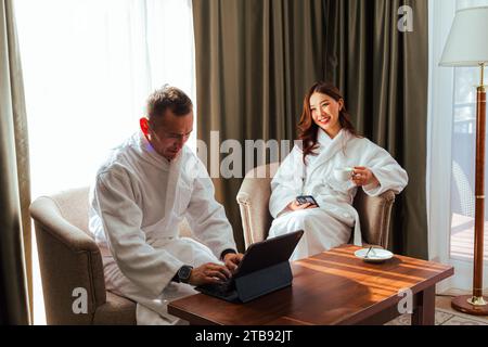 Asiatisches Paar in weißen Bademänteln, das während der Flitterwochen im Hotelzimmer auf einem Stuhl sitzt. Männer, die am Tablet-Computer arbeiten, und Frauen, die Tee aus der Tasse trinken Stockfoto