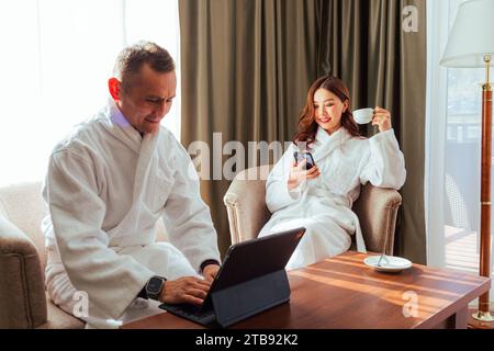 Asiatisches Paar in weißen Bademänteln, das während der Flitterwochen im Hotelzimmer auf einem Stuhl sitzt. Männer, die am Tablet-Computer arbeiten, und Frauen, die Tee aus der Tasse trinken Stockfoto