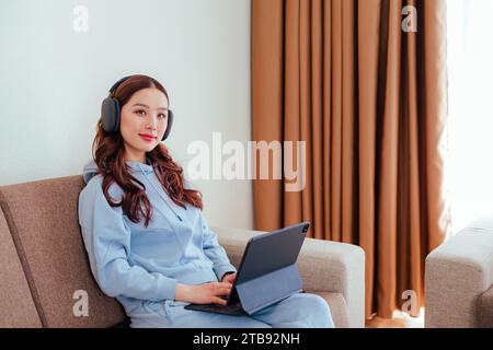 Nahaufnahme einer lächelnden freischaffenden asiatischen Frau, die zu Hause Kopfhörer trägt, einen Tablet-Computer benutzt und die Kamera betrachtet Stockfoto