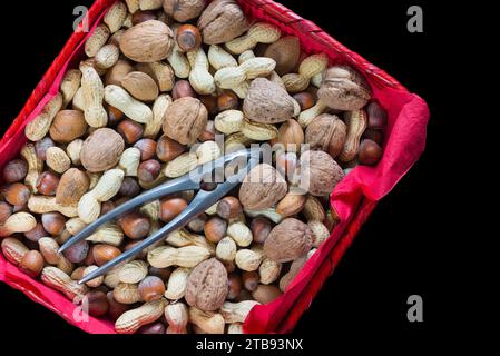 Mischung aus getrockneten Früchten mit Schale in einem roten Korb mit Nussknacker auf schwarzem Hintergrund. Nahaufnahme und Überkopfansicht von Haselnüssen, Walnüssen, Erdnüssen Stockfoto